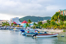 Boats Docked On Tropical Caribbean Island Coast With White Sand And Coconut Palm Trees. Tourists And Locals Shop At Stores And Restaurants On Shore. Mountain Landscape In Background.