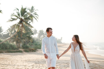 Wall Mural - The bride and groom caucasian couple walk together walk along the beach sand shore. Coconut palm trees on background and ocean seascape. Honeymoon in asia