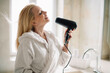 © JustLife - Beautiful woman enjoying in hotel room. Young happy smiling woman wearing bathrobe using hair dryer in bathroom.
