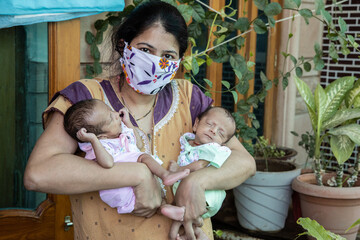 Young asian indian mother wearing mask holding her two newborn twin babies, new normal lifestyle, Stay Home Taking Extra Care Due To new strain of Covid-19 Pandemic. 