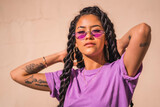 Urban session. Young dark-skinned woman with long braids wearing purple glasses on a plain cream-colored background, with serious look