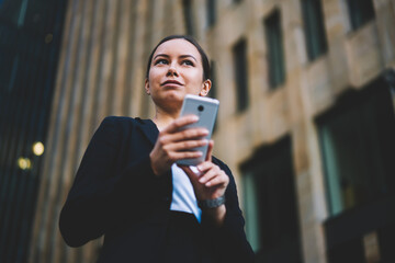 Wall Mural - Below view of successful female professional manager browsing internet websites in searching financial news on modern mobile phone.Businesswoman with telephone in hand looking away standing outdoor