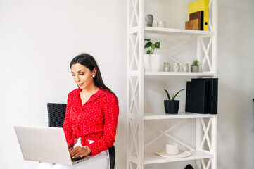 Pretty young woman using laptop for web surfing, online shopping. Girl in red sits on the chair with a laptop indoors