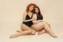 Diversity. Models With Different Figure And Size Portrait. Female Friends Sitting On Floor. Smiling Brunette And Redhead In Black Bodysuits Posing On Beige Background.