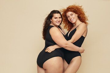 Wall Mural - Diverse. Different Women Portrait. Hugging Female Friends Posing On Beige Background. Smiling Brunette And Redhead In Black Bodysuits.