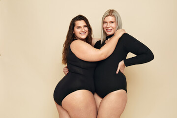 Wall Mural - Plus Size Models. Full-figured Girls Portrait. Brunette And Blonde In Black Bodysuits Posing On Beige Background. Smiling Female Looking At Camera. Body Positive As Lifestyle.