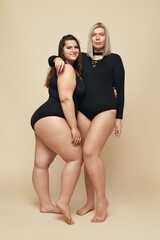 Wall Mural - Plus Size Models. Full-figured Women Full-Length Portrait. Brunette And Blonde In Black Bodysuits Posing On Beige Background. Body Positive Concept.