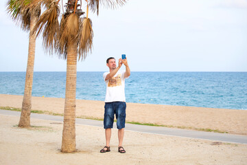 A man in a T-shirt and denim shorts takes a selfie on his smartphone, smiling. A young man communicates via video link with family.Life after quarantine. Video conference. Chat. Palm trees at sunset