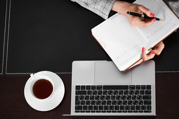  Woman with laptop and notepad. Woman's hands write in a notebook. A cup of tea. Using a computer concept.