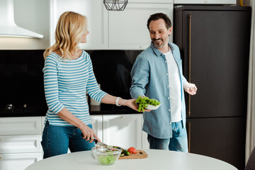 Smiling man giving lettuce to wife cutting vegetables in kitchen