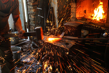 Blacksmith manually forging the molten metal on the anvil with spark fireworks.