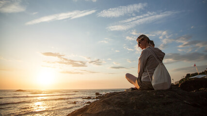 Poster - A young woman sitting on the rocky Atlantic coast during sunset