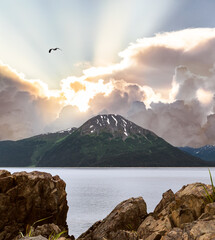 Sunrays in the sky behind the Alaskan mountain on the water with rocks on the shore 