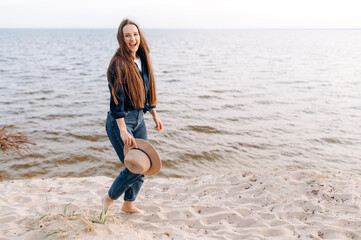 Wall Mural - Happy smiling girl with long hair in casual stylish clothes walks barefoot on the sand by the sea at sunset in a positive mood