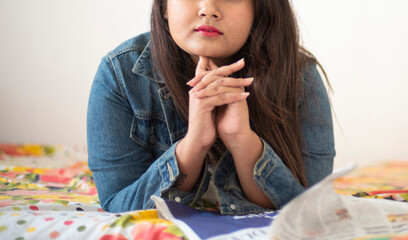 Wall Mural - half face portrait of Indian Bengali beautiful and cute brunette girl in a casual blue jeans shirt is reading newspaper lying on bed in relaxing and casual mood. Indian lifestyle and fashion portrait