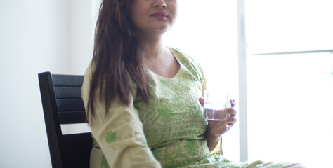 Wall Mural - Indian Bengali beautiful and sexy brunette girl in an ethnic green dress drinking water from a transparent glass sitting in front of a glass door. Indian lifestyle and fashion portrait.