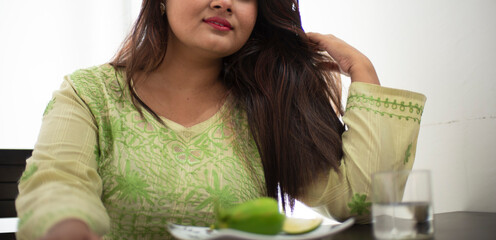 Wall Mural - Low angle half face portrait of an Indian Bengali beautiful and cute brunette girl in ethnic green dress is smiling and talking sitting on a chair. Indian lifestyle and fashion portrait.