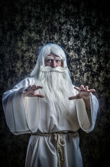 Wall Mural - A grey-haired bearded severe wizard in a white cloak is doing magic against dark-gold background.