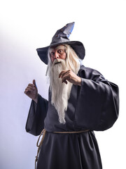 A stern grey-haired bearded wizard in a gray cassock and a cap is practicing sorcery and doing magic against a light background.