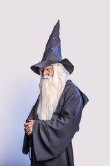 Wall Mural - A severe grey-haired bearded sorcerer in a gray cassock and a cap is practicing sorcery and doing magic against a light background.