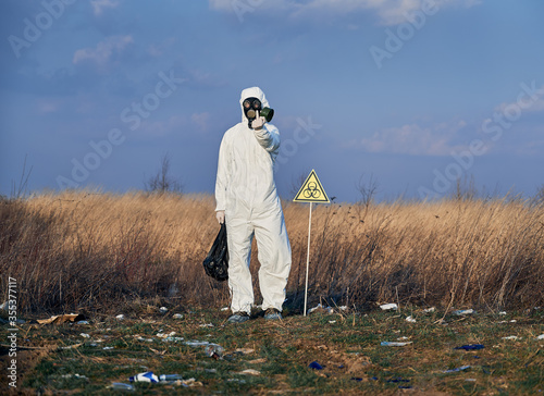 Full length of angry environmentalist in protective suit standing in field with garbage and biohazard sign. Scientist in gas mask doing rude hand gesture while picking up trash. Concept of ecology