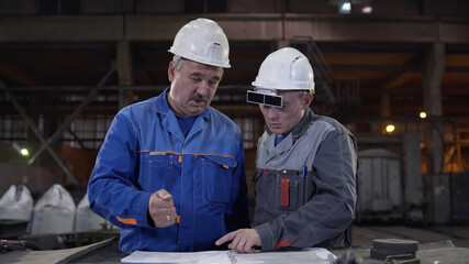 Two workers at heavy industry factory have conversation while looking at blueprint. Engineers explore and discuss project in heavy industry factory. Foundry for Metal Cast and Production.