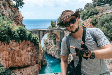 Sticker - The handsome man traveler enjoys a wonderful view of the beach and the stone bridge. Sunglasses and a kangaroo leather cowboy hat. Travel and vacation. Fiordo di Furore, Amalfi, Salerno coast.