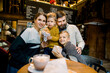 © sofiko14 - Joyful young smiling family with children sitting in cafe at the table, posing to camera with fresh tasty croissants in hands. Family breakfast in cafe