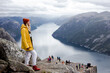 Smiling blonde woman hiker in yellow raincoat stays on the top of Preikestolen mountain (Preacher's Pulpit or Pulpit Rock) with a lot of tourists and enjoys the view of Lysefjord and cliffs background