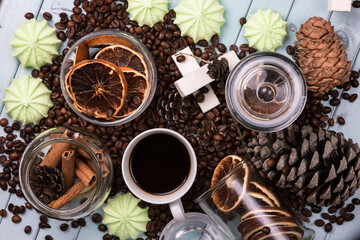 A white Cup of coffee on coffee beans, on a light blue wooden table. Wooden table. Fir cones on the table. Green marshmallows and white marshmallows. Dried citrus. Brown background.