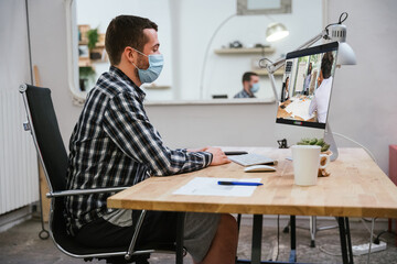 Young millennial in video call with the office, man wearing mask for the other people in the room but he is in casual dress at the computer - Concept of safety in lockdown from Coronaviruss, Covid-19