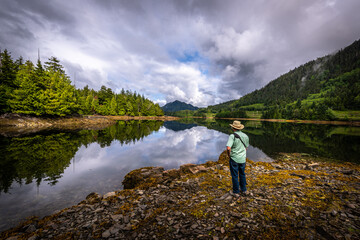 A Man standing at the shore in the morning enjoying the motionless water of the pacific ocean and its mirror like reflection in an inlet near Prince Rupert, British Columbia, Canada.