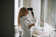 © JustLife - Young happy smiling woman wearing bathrobe and  using hair dryer in bathroom. Beautiful woman enjoying in hotel room.
