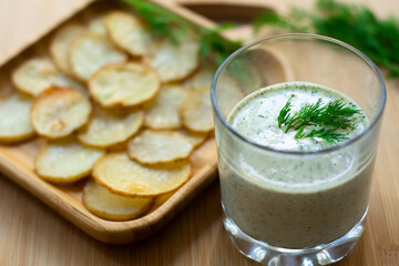 Dill and sour cream dip for homemade potato chips made vegan: healthy snack with dill, tahini, water, vinegar and lemon juice in a glass. Thinly sliced and baked potatoes in the back