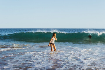 Wall Mural - Young girl in ocean water looking back at approaching wave. Summer fun on a beach.