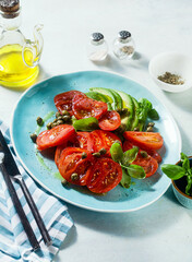 Wall Mural - summer salad with ripe tomatoes and avocado with capers in a blue plate on the table