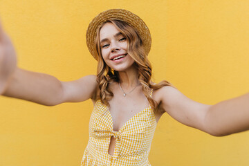 Wall Mural - Debonair european woman in hat making selfie with happy smile. Fascinating girl with wavy hairstyle taking picture of herself on yellow background.