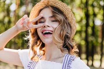 Wall Mural - Close-up outdoor portrait of cheerful girl in straw hat. Enchanting young woman posing in forest with peace sign.