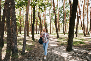 Wall Mural - Full-length portrait of glad girl in jeans walking in forest. Winsome curly woman with backpack posing in park.