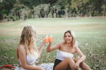 Wall Mural - Happy friends on picnic in park, two young beautiful teen girls having fun and drinking orange juice while spending time together in the garden, woman outdoor friendship concept.