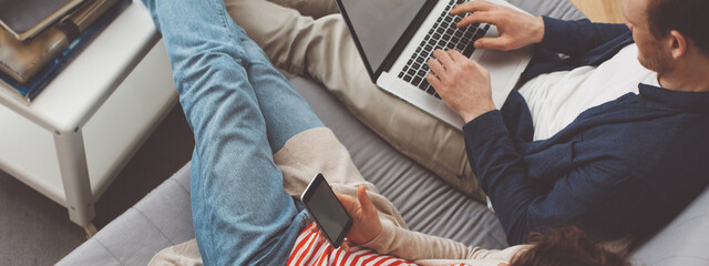 Couple spending time together at home. Man using computer and woman typing message with mobile phone. Sitting together on the sofa. Modern family life