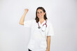 © Irene - Young beautiful woman standing over white isolated background wearing her doctor uniform and doing a gesture with his arm of strength and victory.