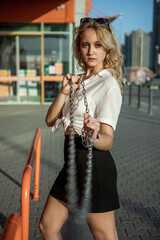 A girl in a white shirt poses against the background of an orange building. The blonde in sunglasses holds iron chains with her hands. Portrait of a girl on the street