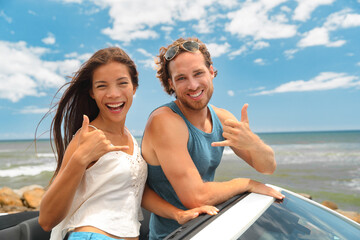 Road trip happy young tourists couple excited in convertible sports car doing shaka sign in happiness. Summer travel vacation in Hawaii.