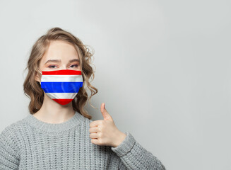 Wall Mural - Happy woman in face mask holding thumb up with Thai flag background. Flu epidemic and virus protection concept