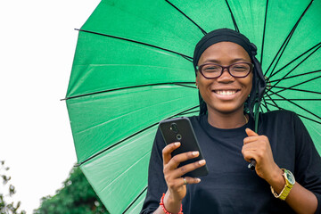 Wall Mural - a young african woman viewing something on a mobile phone and feeling excited, smiling, wearing black, holding an umbrella, looking at camera