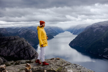 Wall Mural - Blonde woman hiker in yellow raincoat and mustard hat stays on the cliff of Preikestolen mountain (Preacher's Pulpit or Pulpit Rock) with background of Lysefjord and low cloudy sky in Norway