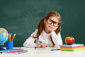 Wall Mural - funny child   schoolgirl  girl student sitting at table near school blackboard