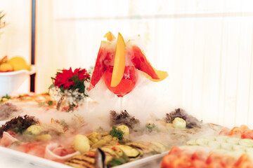 Smoking glass at fruit and vegetable buffet table. Closeup of glass of red beverage with white dense fog. Chemical reaction of dry ice with water.