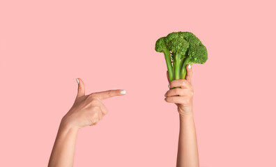 Wall Mural - Woman pointing at broccoli in her hand against pink background, close up view